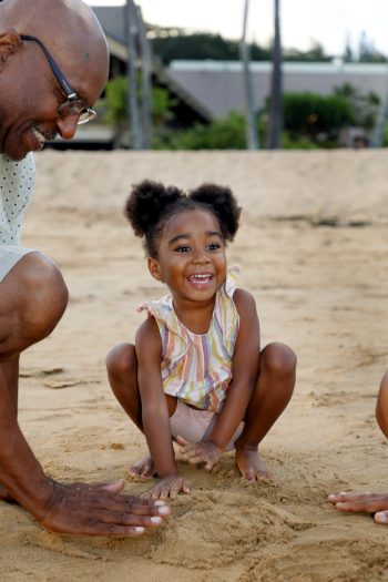 A smiling young girl squats on sandy ground while an adult beside her reaches toward the sand.