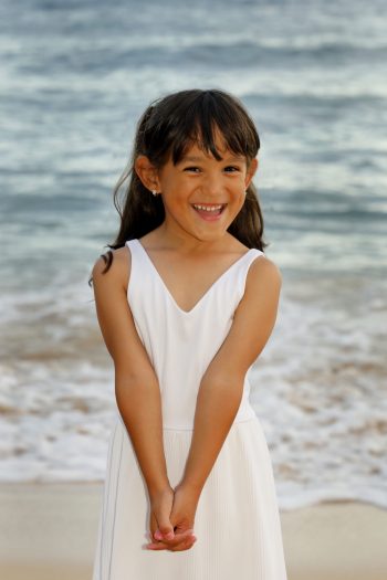 Young girl in a white dress standing on a beach, smiling with her hands clasped in front of her.