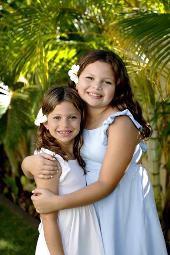 Two girls in light dresses, each with a white flower in their hair, smiling and hugging outdoors among palm leaves.