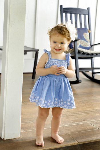 Toddler in a blue dress with white embroidery stands barefoot indoors near a pillar, smiling at the camera.
