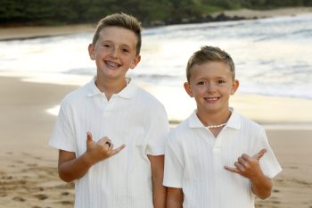 Two boys in white shirts stand on a beach, both smiling and making a shaka hand gesture.