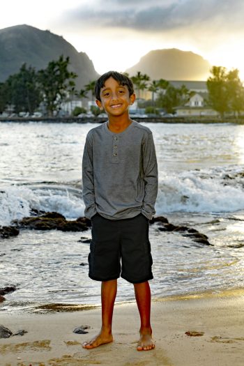 Boy stands barefoot on a sandy beach with waves, trees, and mountains in the background at sunset.