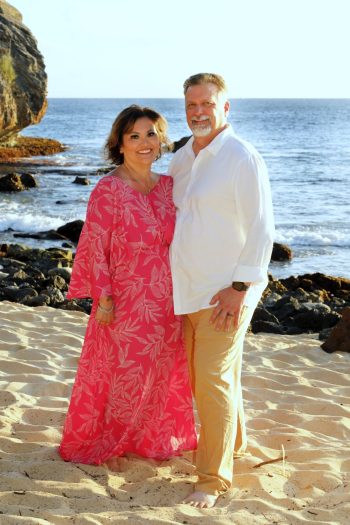 A couple stands together on a sandy beach, with the ocean and rocky shoreline in the background.