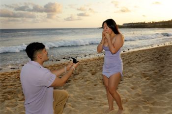 A man kneels and proposes with a ring to a surprised woman on a sandy beach at sunset.