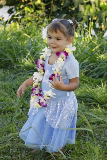 Young child in a blue dress and flower lei stands barefoot on grass, with greenery and sunlight in the background.
