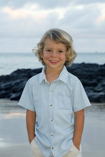 Young child with blonde hair in a light denim shirt stands on a beach with rocks and ocean in the background.