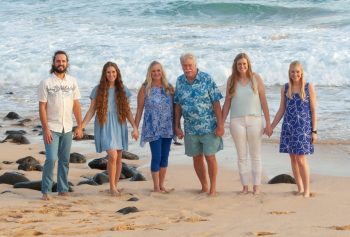 Six people stand barefoot on a beach, holding hands in a line with ocean waves in the background.