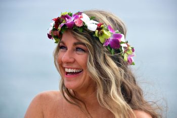 Smiling woman with long wavy hair wearing a colorful floral crown, with a blurred outdoor background.