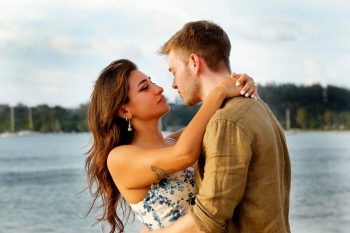 A couple stands by the water, embracing and gazing into each other's eyes on a cloudy day.