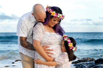 A man, woman, and child stand on a beach. The woman is pregnant and all three wear floral crowns and embrace her belly.