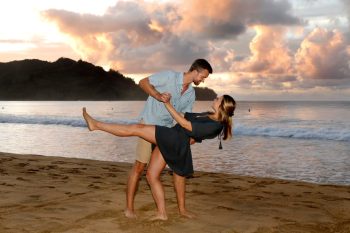 A man dips a woman while standing on a sandy beach at sunset with waves and mountains in the background.