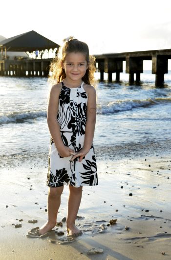 A young girl stands barefoot on a sandy beach with a pier and pavilion in the background at sunset.