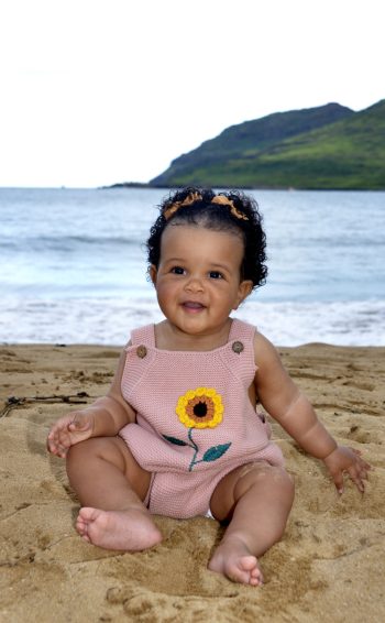 A smiling baby in a pink romper with a sunflower sits on sandy beach with ocean and green hills in the background.