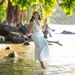 A woman in a white dress and flower lei sits on a wooden swing over shallow water under leafy branches.