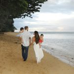 A couple walks along a sandy beach with two young children, one parent carrying each child near the shoreline.