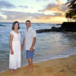A man and woman stand barefoot on a sandy beach at sunset with waves and palm trees in the background.