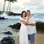 A couple stands barefoot on a sandy beach, embracing, with ocean waves and palm trees in the background.
