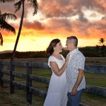 Couple standing by a wooden fence, smiling at each other during sunset with palm trees in the background.