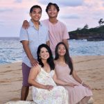 Four people pose together on a sandy beach at sunset, with the ocean and a rocky shore in the background.