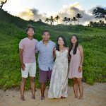 Four people stand barefoot on a sandy beach with greenery and trees in the background at sunset.