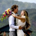 A couple in wedding attire embraces outdoors, smiling at each other, with mountains and greenery in the background.