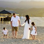A family of four walks barefoot on a sandy beach near a pier at sunset, holding hands and dressed in white.