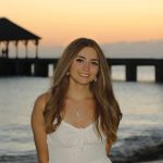 Young woman in a white dress smiles on a beach at sunset, with a pier and pavilion in the background.