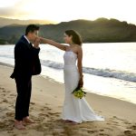 A couple in wedding attire stands barefoot on a beach at sunset, with the groom kissing the bride’s hand.