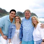 Four adults standing close together on a beach with waves and greenery in the background, all smiling at the camera.
