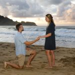 A man kneels and holds a woman's hand on a sandy beach at sunset, with waves and clouds in the background.