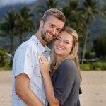 A smiling couple embraces on a sandy beach with palm trees and mountains in the background.