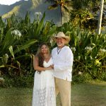 A smiling couple in white outfits stands on grass with tropical plants and mountains in the background.