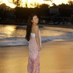 Woman in a pink and white dress stands barefoot on a beach at sunset, looking back and smiling at the camera.