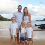 A family of five poses barefoot on a sandy beach with water and greenery in the background.