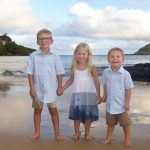 Three young children stand barefoot on a beach, holding hands and smiling, with the ocean and hills in the background.