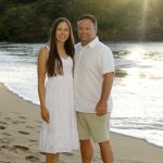 A woman and a man stand barefoot on a sandy beach near the water, smiling at the camera with greenery in the background.