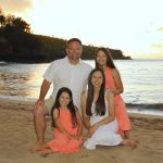 A family of four poses on a sandy beach at sunset, with the ocean and trees in the background.