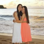 Two people in summer dresses stand barefoot on the beach, hugging and smiling at sunset with waves in the background.