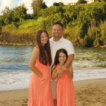 A man stands on the beach with two young girls in matching coral dresses, with greenery and water in the background.