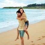 A man gives a woman a piggyback ride on a sandy beach near the ocean, both smiling and barefoot.