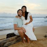 A couple poses and smiles on a sandy beach, with waves, driftwood, and greenery in the background.