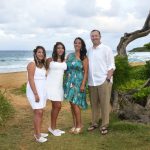 Four people stand together on a grassy area near a beach with ocean, sand, and trees in the background.