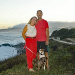 A smiling couple stands on a grassy cliff with two dogs, overlooking the ocean at sunset.