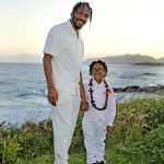 An adult and a child dressed in white stand on grass by the ocean, smiling at the camera at sunset.