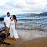 A couple stands barefoot on a sandy beach near driftwood, facing each other with the ocean and mountains in the background.