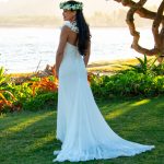 A woman in a white dress and flower crown stands on grass near the ocean, surrounded by tropical plants and trees.