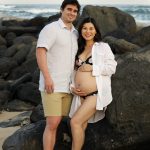 A couple stands barefoot on a rocky beach, with the woman in a bikini and open shirt, holding her pregnant belly.