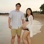 A man and a pregnant woman stand barefoot on a sandy beach near the water, smiling at the camera.