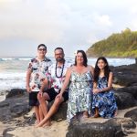 A family of four sits on black rocks by the beach, with waves and greenery in the background under a partly cloudy sky.
