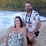 A woman and man in tropical attire pose together on a sandy beach with trees and ocean in the background.